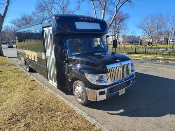 A black bus labeled "INTERNATIONAL 32 PASSENGER BUS" is parked on the side of a road with trees and a grassy area in the background on a clear day.