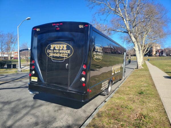 A black INTERNATIONAL 32 PASSENGER BUS with "FUI CALL FOR SERVICE" and a phone number printed on the rear is parked on the side of a street next to a sidewalk and trees.