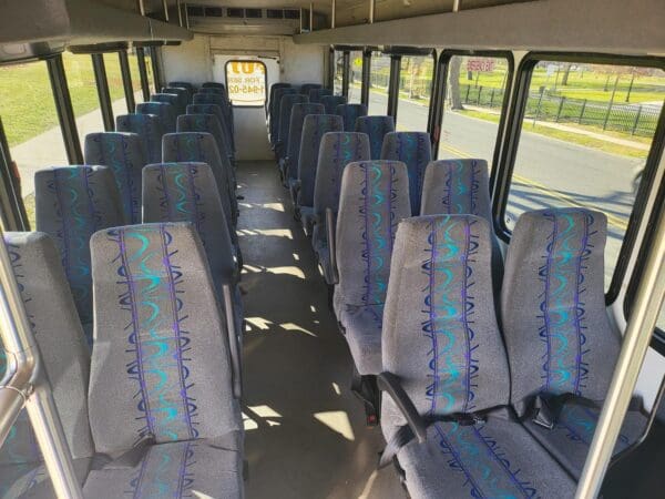 Interior of an empty INTERNATIONAL 32 PASSENGER BUS with patterned seats, looking forward towards the driver's area. Sunlight is streaming through the windows on the right side.