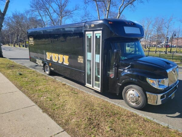 A black INTERNATIONAL 32 PASSENGER BUS with the FUJI logo is parked on the side of the street near a grassy area and trees under a clear blue sky.