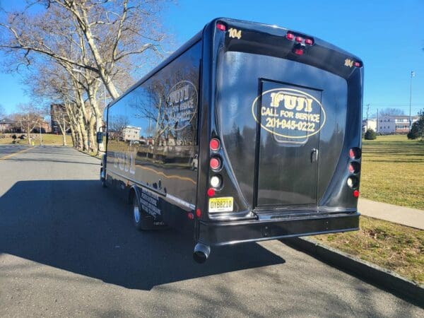 A black INTERNATIONAL 32 PASSENGER BUS with "Fiji" branding and "Call for Service" text on the back, parked on a road near grassy areas and trees. The contact number 201-845-0222 is displayed on the rear window.