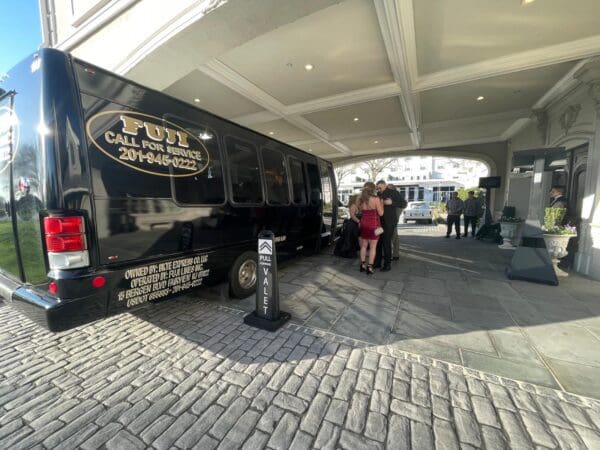 A small group of people stands near a black shuttle bus parked under a portico outside a building, with a "Valet" sign placed nearby.